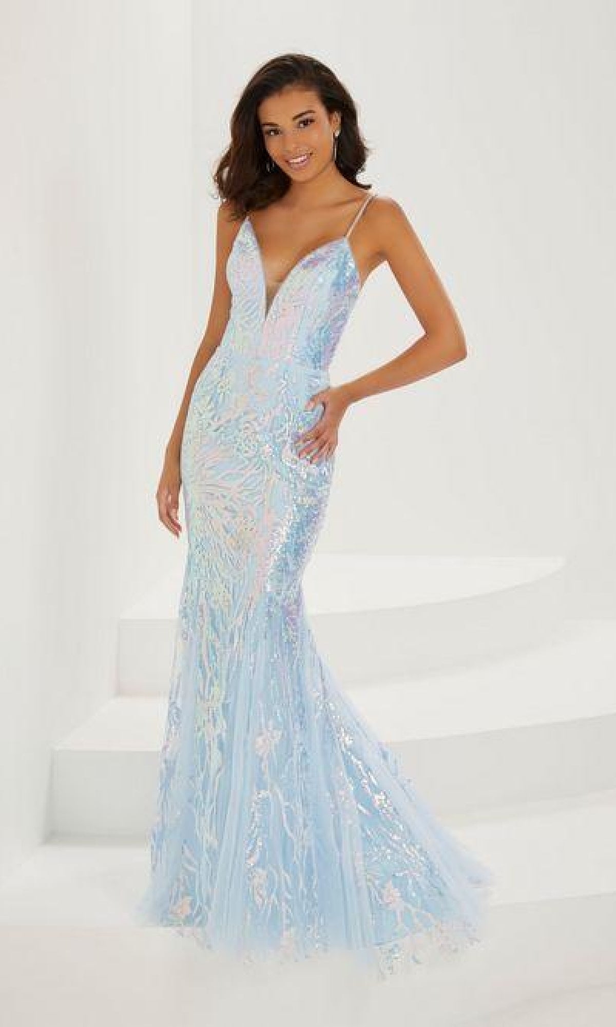 A woman stands on white stairs against a plain background, wearing the Long Sequin Prom Dress: Tiffany 16944—a light blue, iridescent, floor-length gown with a tulle overlay, thin straps, and a deep V neckline.
